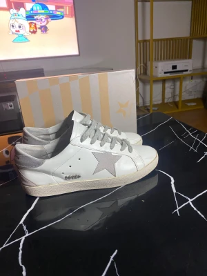 GGDB Superstar vita sneakers stl 43 - Säljer ett par GGDB Superstar sneakers i vitt skinn med grå snörning och en grå stjärna på sidan. Skorna har rund tå, platt sula och silverfärgad häl. Klassisk och clean design med box medföljer. Perfekt för dig som gillar streetstyle och exklusiva märken.