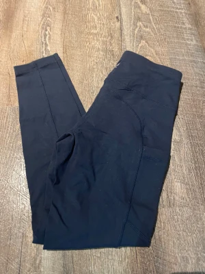Svarta leggings från SOC med snörning - Säljer ett par svarta leggings från SOC i storlek S. De har praktisk snörning i midjan och en ficka på sidan. Materialet är stretchigt syntet som andas och passar perfekt för träning. Snygg och enkel design som är lätt att matcha med din träningsoutfit.