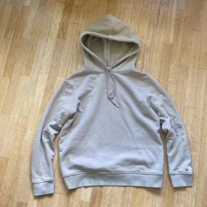 Sällsynt Tommy hilfiger hoodie  - Snygg beige hoodie från Tommy Hilfiger med broderad logga på ärmen och klassisk huva med snörning. Tröjan har långa ärmar, ribbade muddar och en relaxed passform. Perfekt för dig som gillar stilrena och bekväma plagg. Säljer pga att den är för liten