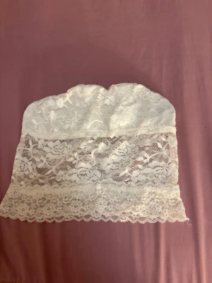 Vit spets bandeau tubtopp - Säljer en vit bandeau tubtopp i spets med blommigt mönster. Toppen är axelbandslös och har en romantisk vibe med genombruten spets och fina detaljer längs nederkanten. Perfekt för att styla med jeans eller kjol för en cool look.