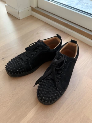 Svarta Christian Louboutin sneakers med nitar - Säljer ett par svarta sneakers från Christian Louboutin i sammet med coola svarta nitar på tån och klassisk röd sula. Skorna har snörning och en låg profil, perfekt för dig som gillar edgy designerstil. Insidan är beige och de har rund tå.