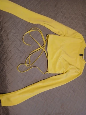 Gul croppad långärmad topp från DIVIDED - Säljer en ljusgul croppad topp från DIVIDED i storlek XS. Toppen har långa ärmar och coola snörningsdetaljer i midjan som kan knytas på olika sätt. Materialet är mjukt och stretchigt, troligen polyester. Perfekt för dig som gillar färg och trendiga detaljer.