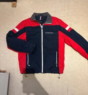 Peak Performance pufferjacka röd/blå L - Peak Performance pufferjacka i storlek L med snygg färgblockad design i rött och mörkblått. Jackan har vit dragkedja, ståkrage i grått, logga på bröstet och stor logotyp på ryggen. Två fickor med dragkedja och justerbar nederkant med snörning.