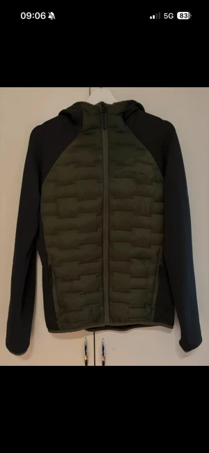 Peak Performance grön pufferjacka - Snygg Hybrid från Peak Performance i mörkgrönt och svart med huva och hel dragkedja. Jackan har quiltad front och rygg, samt mörka ärmar och sidopaneler. Perfekt för kyliga dagar och har en sportig, modern look.