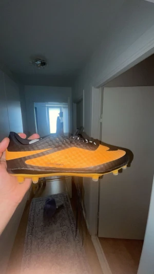 Nike Hypervenom fotbollsskor orange/svart - Nike Hypervenom fotbollsskor i en snygg orange och svart färg med mönstrad ovandel och klassisk Nike-logga på sidan. Skorna har snörning och platt sula med fasta dobbar för gräsplan. Lätta och smidiga med syntetmaterial som ger bra känsla på bollen.
