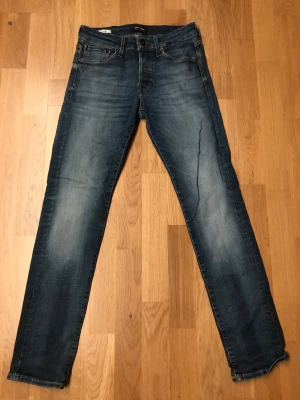 Slim Glenn jeans från Jack & Jones - Snygga blå jeans från Jack & Jones, modell Slim Glenn. De har en slim passform, klassisk femficksdesign och snygga slitningar på benen. Jeansen är tillverkade i jeansmaterial och har en mörkblå tvätt med ljusare partier för en cool look. Är ett litet hål på ena benet, men är igen sytt så syns knappast det syns på en utan bilderna, 