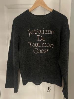 Mörkgrå stickad tröja med broderi - HELT NY Mysig mörkgrå stickad tröja med ljusrosa broderad text 'Je t'aime De Tout mon Coeur' på framsidan. Tröjan har rund hals och långa ärmar, perfekt för att ge din outfit en personlig touch.