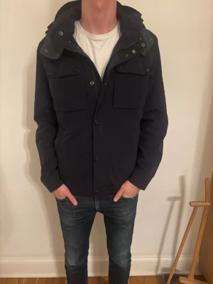 Mörkblå vindjacka Tommy Hilfiger M - ⭐️⭐️⭐️Snygg mörkblå jacka från Tommy Hilfiger i storlek M. Jackan har en bortagbar luva och snygg diskret logga på ärmen. Liten defekt på insidan av jackan men påverkar inte. Riktigt snygg och passar perfekt med en tröja till årets vinter. ⭐️⭐️⭐️