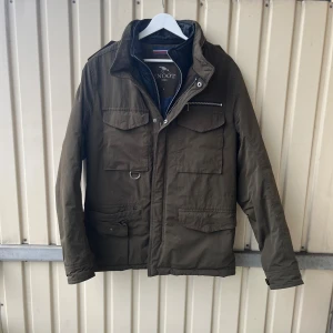 Olivgrön FieldJacket från Snoot - Säljer en olivgrön parkajacka från Snoot med flera praktiska fickor, dragkedja och knappar framtill. Jackan har hög krage, innerfoder i mörkblått och coola detaljer som metallring och bröstficka med dragkedja. Perfekt för dig som gillar funktionell och snygg stil.