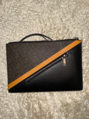 Fendi svart och brun  - Snygg kuvertväska från Fendi i svart och brun skinn med klassiskt Fendi-mönster och en diagonal gul detalj på framsidan. Väskan har en silverfärgad dragkedja och ett litet handtag upptill. Perfekt för att bära det viktigaste med stil.