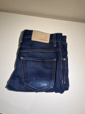 Mörkblå jeans från J.Lindeberg 29/32 - Snygga mörkblå jeans från J.Lindeberg med klassisk femficksdesign och kontrastsömmar. Jeansen har en rak passform och är tillverkade i slitstarkt denim med lätt tvättade detaljer på bakfickan. Perfekt för dig som gillar stilrena och tidlösa jeans.