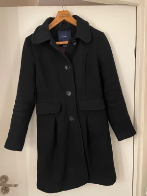 Svart kappa från GANT i ull - Svart kappa från GANT med klassisk krage och knappar framtill. Jackan har fickor med lock och snygga veck nedtill som ger en stilren siluett. Materialet är 80% ull och 20% polyamid, vilket gör den både varm och mjuk. Perfekt för dig som gillar tidlös design.