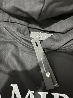 Svart AMIRI hoodie med vit logga - Svart hoodie från AMIRI med stor vit logga och grafiskt tryck på bröstet. Tröjan har huva, känguruficka och ribbade muddar vid ärmslut och nederkant. Materialet är mjuk bomull, perfekt för chill dagar. Snygg och enkel design som sticker ut.