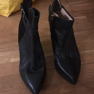 Svarta kilklacksstövletter i skinn - Snygga svarta stövletter med kilklack och spetsig tå. Skorna är gjorda i skinn och har en dekorativ rem med spänne runt ankeln samt dragkedja på insidan. Perfekta för dig som gillar en edgy och stilren look.