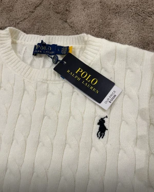 Vit stickad tröja från Polo Ralph Lauren - Vit kabelstickad tröja från Polo Ralph Lauren med klassisk rund hals och långärmad design. Tröjan har den ikoniska svarta Polo-loggan broderad på bröstet och ribbade muddar vid ärmslut och nederkant. Perfekt för dig som gillar stilren och tidlös look.