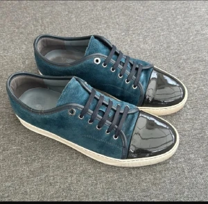 Blå mockasneakers från Lanvin - Snygga sneakers från Lanvin i mörkblå mocka med svart lackad rund tå och svarta snören. Skorna har vit platt sula och metallfärgade öljetter. Perfekt för dig som gillar exklusiv och unik stil med coola materialmixar.