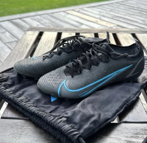 Nike Mercurial vapor 14 elite ag - Säljer ett par Nike Mercurial fotbollsskor i svart med blå detaljer och snörning. Sälj då dem är för små! Skorna är i använt skick men har många timmar kvar i sig! Bagen ingår, Skriv vid fler funderingar:) 