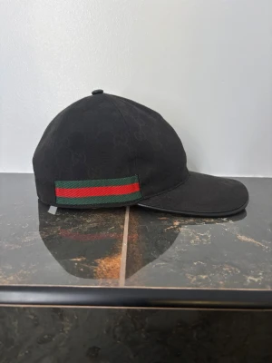 Svart Gucci canvas - Snygg svart keps från Gucci med diskret GG-monogram över hela ytan. På sidan finns en klassisk grön och röd rand. Justerbar rem baktill i skinn och insida med Gucci-etikett. Perfekt för dig som vill ha en lyxig streetwear-look.