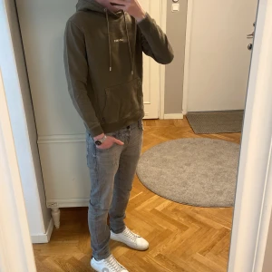 grön hoodie från Les Deux - Säljer en olivgrön hoodie från Les Deux med vit logga på bröstet. Tröjan har huva med snörning, magficka och ribbade muddar. Perfekt för chill dagar och enkel att matcha med jeans eller joggers. Materialet är mjuk bomull som känns skönt mot huden. Modellen är 185 lång och väger 70 kg. Skriv vid minsta fundering.