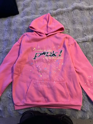 Sp5der P*NK Hoodie Rosa Spider Worldwide Tröja (Storlek M) - Snygg rosa hoodie med huva och magficka. Framsidan har ett coolt tryck med texten 'nevermind the spider, heres the slime', vita prickar och ett spindelnätsmönster med färgglada stjärnor. Mjuk insida och ribbade muddar. Perfekt för dig som gillar statement-plagg.