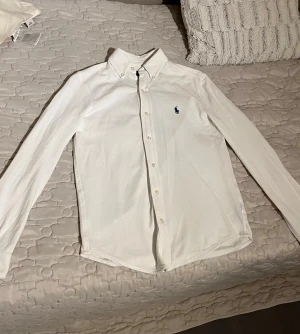 Vit skjorta från Ralph Lauren - Klassisk vit skjorta från Ralph Lauren i lätt mesh-bomull. Passar S också. Skjortan har button down-krage, långa ärmar och knappar framtill. Ikoniska blå Polo-loggan broderad på bröstet. Perfekt passform och stilren look.