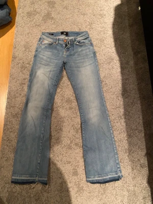 Blå bootcut jeans från Roxy - Snygga ljusblå jeans från Lbt i modell Roxy med klassisk femficksdesign och bootcut-ben. Jeansen har slitna detaljer vid bensluten och kontrastsömmar. Passar dig som gillar en avslappnad och tidlös look. Perfekt till sneakers eller boots.