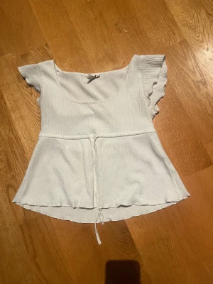 Vit blus med volangärmar och knyt - Supersöt vit blus med volangärmar och rundad halsringning. Toppen har en ribbad struktur och ett knytband i midjan som ger en snygg form. Materialet känns mjukt och luftigt.