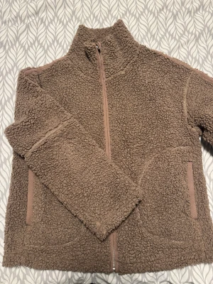 Brun teddyjacka från Nelly - Mysig brun teddyjacka från Nelly i storlek S. Jackan har hög krage, två stora fickor framtill och stängs med dragkedja. Materialet är fluffigt och ger en cool, avslappnad vibe. Perfekt för kyliga dagar när du vill vara både varm och snygg.