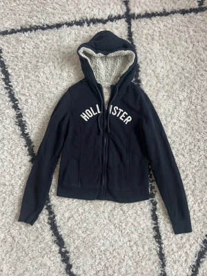 Mörkblå teddyhoodie från Hollister - Mysig mörkblå/svart hoodie från Hollister med vit broderad logga över bröstet. Den har huva med fluffigt teddyfoder, dragkedja framtill och långa ärmar. Perfekt för kyliga dagar när du vill vara både varm och snygg. Storlek M men upplever den lite mindre i storlek 💙