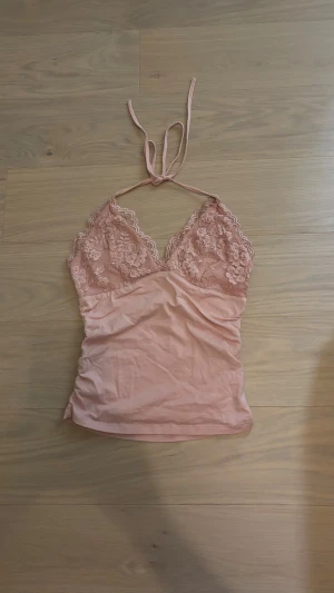 Rosa spetsdetaljerad linnetopp - Supersöt ljusrosa linnetopp med spetsdetaljer och blommigt mönster över bysten. Toppen har smala axelband som knyts i nacken och en omlottkänsla framtill. Materialet är mjukt och stretchigt, perfekt för varma dagar eller när du vill känna dig extra fin.
