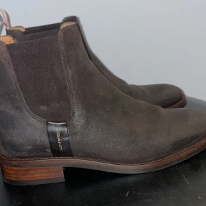 Bruna chelsea boots från Gant - Snygga bruna chelsea boots från Gant i mocka med elastiska paneler på sidorna och träfärgad låg klack. Klassisk rund tå och diskret Gant-logga på sidan. Perfekt för dig som gillar stilrena och tidlösa skor med en modern touch.