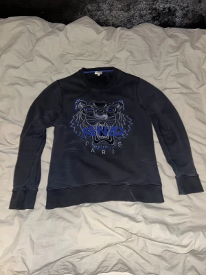 Svart Kenzo sweatshirt  - Svart sweatshirt från Kenzo med ikonisk broderad tiger i blått och grått på bröstet. Tröjan har rund hals, långa ärmar och ribbade muddar. Perfekt för dig som gillar streetstyle och vill sticka ut med ett coolt motiv.