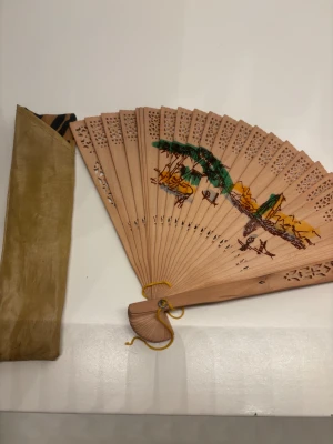 Handmålad solfjäder med fodral - Snygg handmålad solfjäder i trä med utskurna detaljer och motiv i grönt, gult och brunt. Kommer med ett beige fodral i tyg. Perfekt accessoar för varma dagar eller som dekoration.