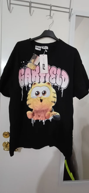Garfield t-shirt från FB Sister XL - Svart t-shirt med loose fit från FB Sister, officiell Garfield Movie-design. Stor tryckt Garfield-figur framtill och rosa text med cool drip-effekt. Mjuk bomullskvalitet och klassisk rund hals. Perfekt för dig som älskar roliga prints och popkultur.