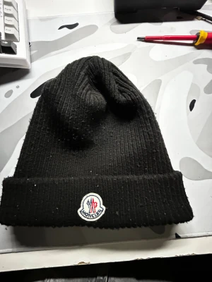 Svart Moncler mössa - Svart ribbstickad mössa från Moncler med uppvikt kant och klassisk Moncler-logga framtill. Snygg och enkel design som funkar till de flesta stilar. Materialet är stickat och mössan har en skön passform som håller dig varm under kyliga dagar.