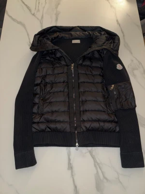 Svart Moncler pufferjacka med huva - Storleken är S-M. Svart pufferjacka från Moncler med stickade ärmar och quiltad front. Jackan har huva, dragkedja framtill och en ficka med dragkedja på ärmen. Ikonisk Moncler-logga på vänster ärm. Perfekt för dig som gillar stilren och sportig look. 