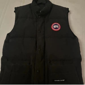 Svart dunväst från Canada Goose - Säljer en svart dunväst från Canada Goose med knappar och broderad logotyp på bröstet. Västen har en klassisk design och är perfekt för kyligare väder.