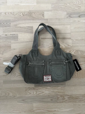 Grå axelväska True Religion🩶 - Snygg grå axelväska från True Religion i jeansmaterial med fickdetaljer och silverfärgade dragkedjor framtill. Väskan har breda handtag och en avtagbar axelrem. Helt ny med tag🩶