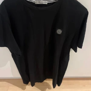 Svart t-shirt från Stone Island - Svart t-shirt från Stone Island med klassisk rund hals och korta ärmar. Diskret logopatch på bröstet och tryckt logga i nacken. Tillverkad i mjuk bomull för skön känsla. Perfekt för dig som gillar stilrena och ikoniska plagg.