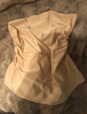 Champagnefärgad tubtopp i satin - Säljer en snygg champagnefärgad tubtopp i glansigt satinmaterial. Toppen är axelbandslös och har en mjuk, draperad form som ger en cool och trendig vibe. Perfekt för dig som vill sticka ut med en stilren och chic look.