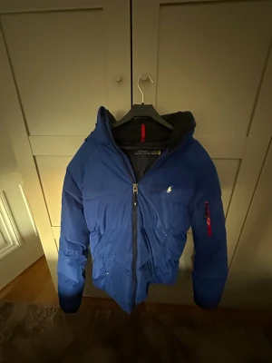 Blå pufferjacka från Polo Ralph Lauren - Blå pufferjacka från Polo Ralph Lauren med huva och dragkedja framtill. Jackan har en röd detalj på insidan av kragen och en liten vit logga på bröstet. Flera fickor, inklusive en med röd dragkedja på ärmen. Perfekt för kalla dagar.Liten fläck vid bröstet men går bort vid tvätt eller skrubbning. Skriv vid minsta fundering😇ny pris 5500.