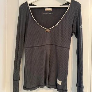 Mörkgrå ribbad topp Odd Molly - Säljer en mörkgrå ribbad topp från Odd Molly med lång ärm och v-ringad hals. Toppen har en dekorativ spetskant och en liten rosett framtill. Märkespatch nertill och broderad detalj på ärmen. Perfekt för dig som gillar snygga detaljer och skön passform.