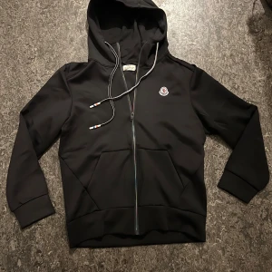 Svart hoodie från Moncler med dragkedja - Svart hoodie från Moncler med huva och snörning, dragkedja framtill och klassisk Moncler-logga på bröstet. Tröjan har långa ärmar och praktisk känguruficka. Snygga detaljer på snörändarna i rött, vitt och blått. Perfekt för chill dagar.