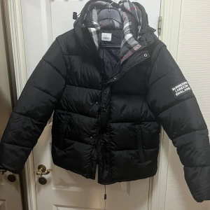 Svart pufferjacka från Burberry - Svart pufferjacka från Burberry London England med avtagbara ärmar så den kan användas som väst. Jackan har huva med klassiskt rutigt Burberry-foder i grått, vitt och rött. Dragkedja och knappar framtill samt två fickor med dragkedja.