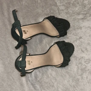 gröna klackskor från H&M - Snygga gröna klackar från H&M. Skorna har en elegant rem runt ankeln med spänne i guld och en mjuk mockaliknande yta. Framtill finns en fin volangdetalj som ger extra stilpoäng. Perfekta för dig som gillar trendiga och bekväma klackar.