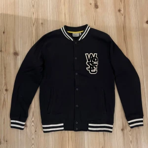 Svart Collegejacka från WESC, strl.S - Svart collegejacka från WESC med vita ränder på krage, ärmslut och nederkant. Jackan har tryckknappar framtill, två fickor och ett stort WESC-märke på bröstet. Tillverkad i mjuk bomull, perfekt för en chill streetwear-look.