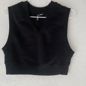 Svart croppad väst från H&M Divided - Svart croppad väst från H&M Divided i storlek XS. Västen har v-ringad hals och breda ribbade kanter. Tillverkad i mjuk bomull och polyester, perfekt att styla över en t-shirt eller skjorta för en cool look.