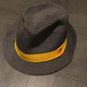 Svart hatt med gultt band från Pählsons - Cool svart hatt i flätad struktur med brett gult band runt kronan och liten etikett med text. Tillverkad i 100% papper, perfekt för att sticka ut i mängden. Klassisk form med nedvikt brätte och markerad topp. 