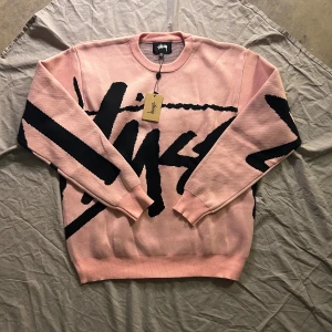 Rosa Stüssy knitted tröja med svart print  - Säljer en rosa Stüssy tröja i storlek XL med stort svart grafiskt print över hela framsidan och ärmarna. Tröjan är tillverkad i 100% bomull och har rund hals samt ribbade muddar. Perfekt för dig som gillar streetwear och vill sticka ut.