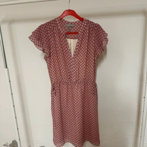 Rödvit mönstrad kortklänning H&M - Snygg kortklänning från H&M i rödvitt geometriskt mönster. Klänningen har volangärmar, v-ringad hals och resår i midjan för en skön passform. Materialet är lätt och luftigt, perfekt för varmare dagar. Passar dig som gillar färg och mönster!